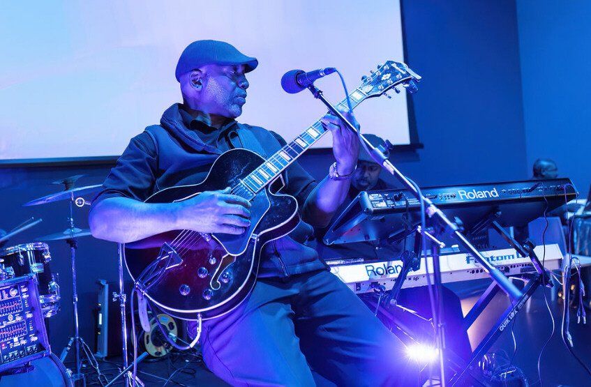Guitarist playing onstage under blue stage lights, with a microphone and keyboard in the background.
