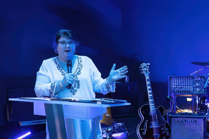 Woman speaking at a lectern on stage, holding a microphone. Blue lighting. Guitar and sound equipment visible.