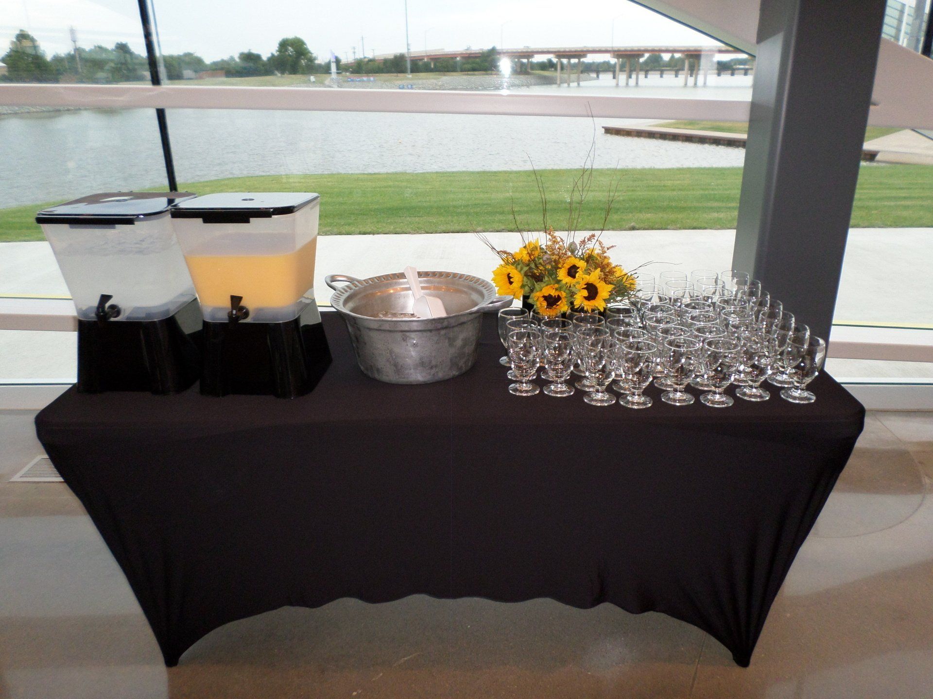 Beverage station with juice dispensers, ice bucket, glasses, and sunflowers on a black tablecloth near a window.