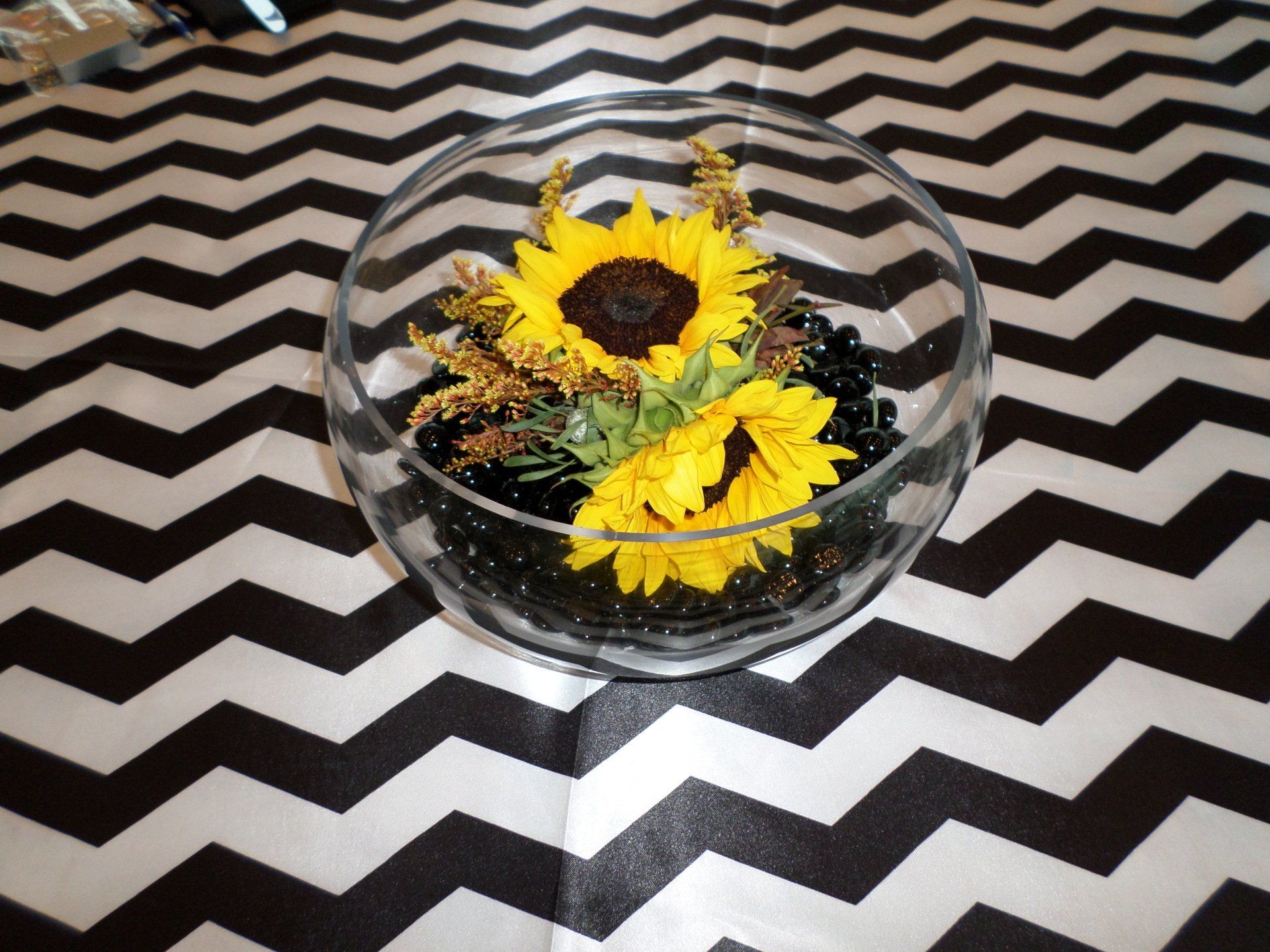 Clear glass bowl with sunflowers and black decorative stones on black and white chevron patterned surface.