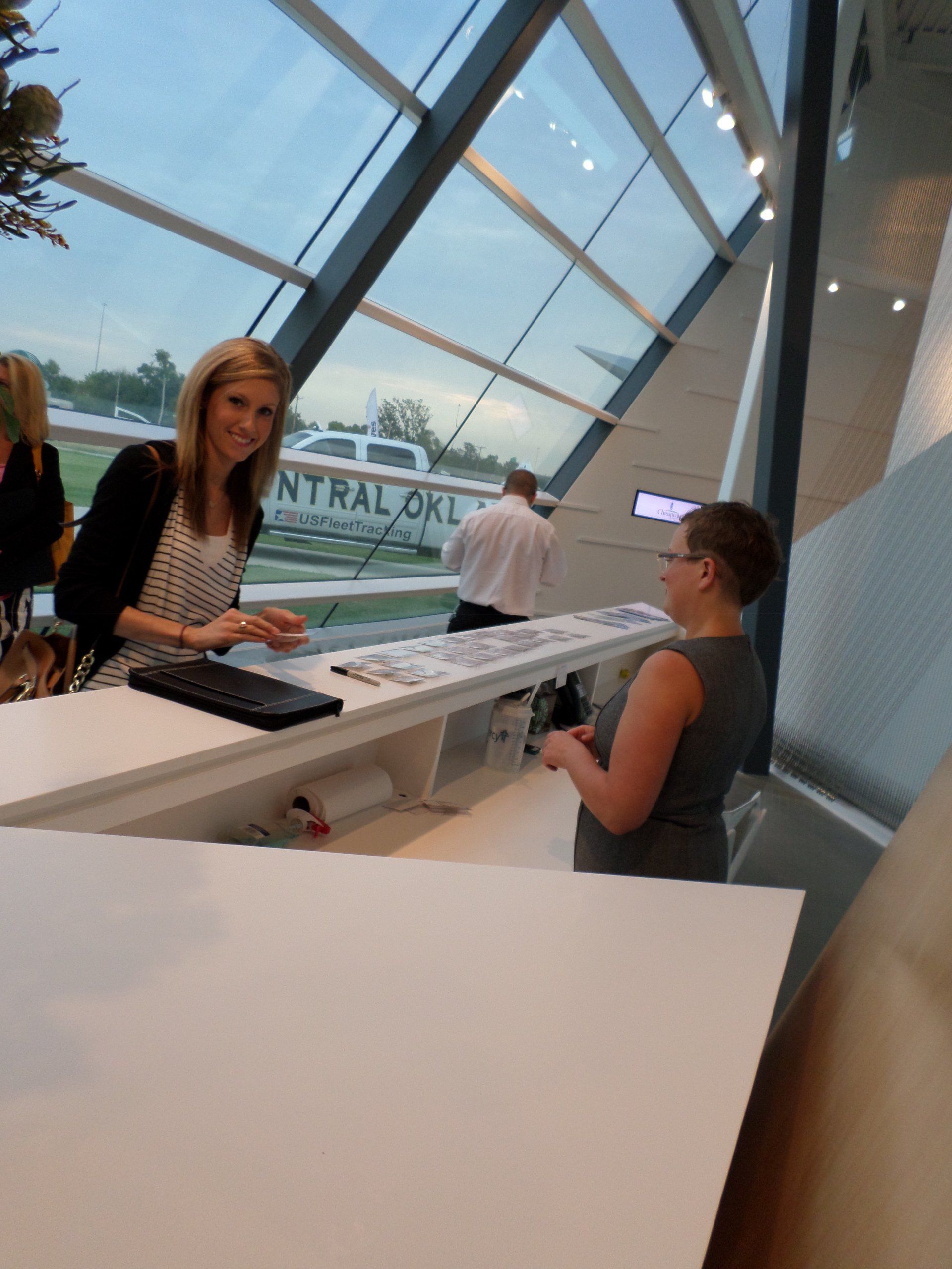 Woman at reception desk smiling at camera. Modern building with large windows.