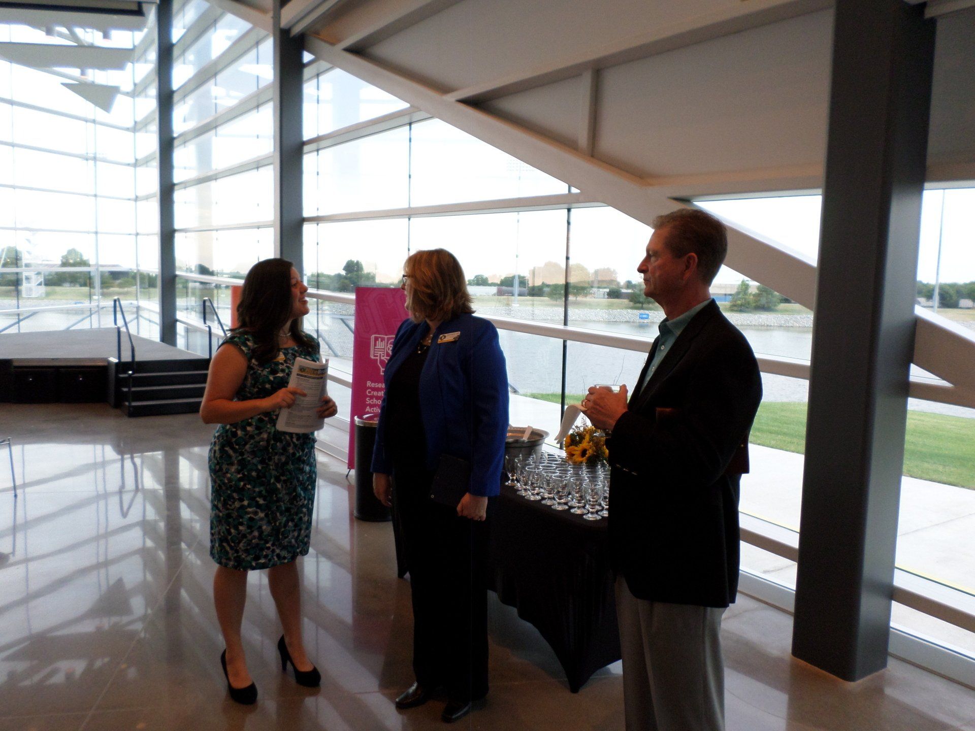 Three people in a modern lobby with large windows. A woman in a dress speaks to a woman in a blue blazer as a man applauds.