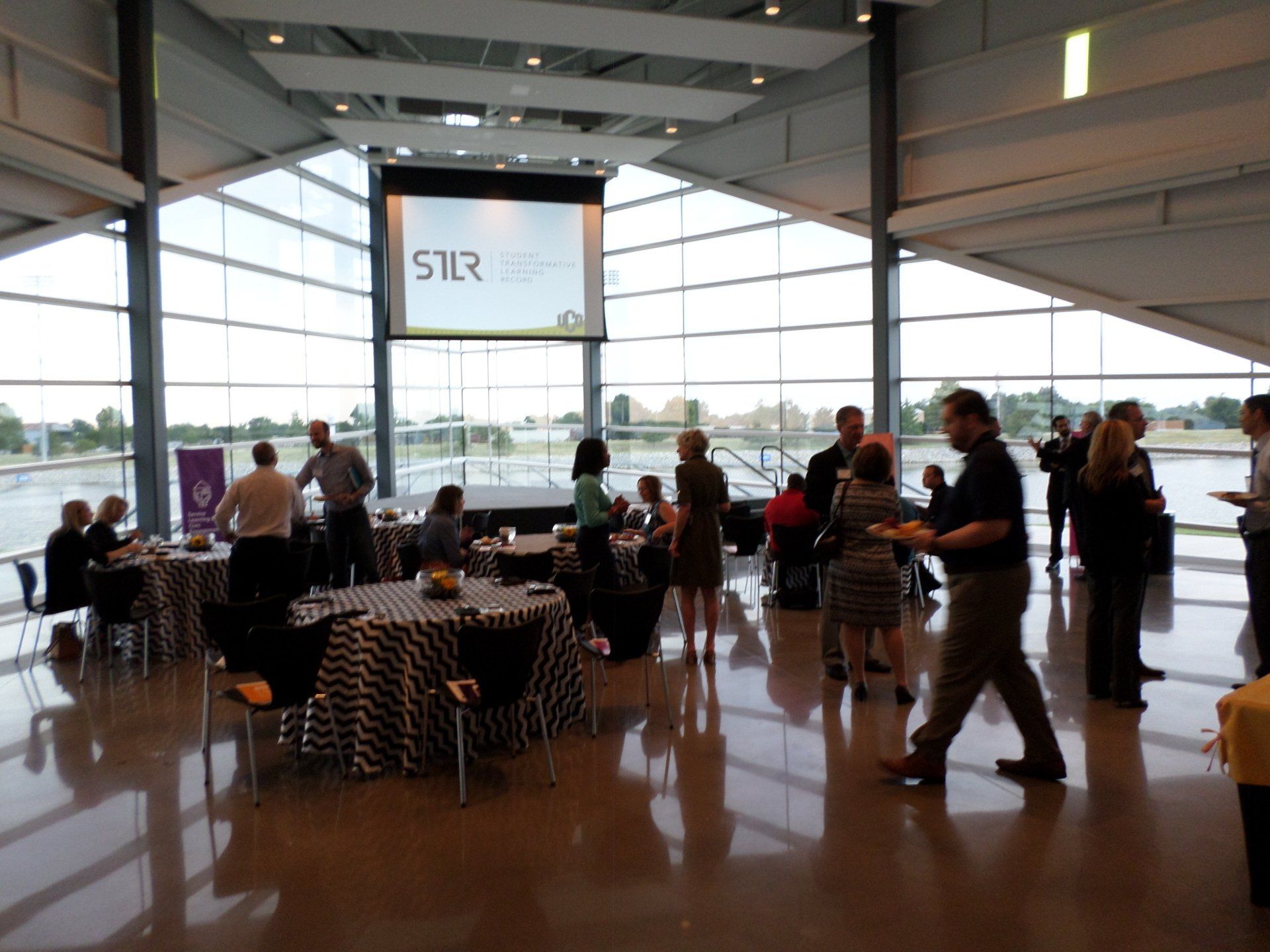 Event reception in a spacious room with tables, people mingling, and a screen displaying a logo.