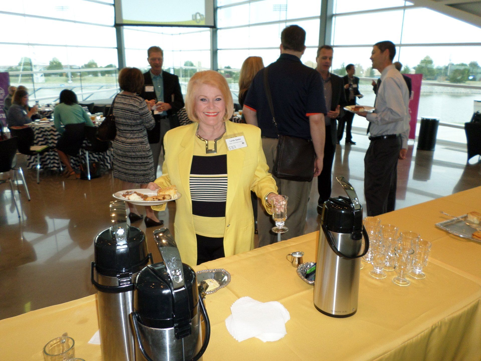 Woman in yellow blazer at a networking event, holding food and drink, smiling.