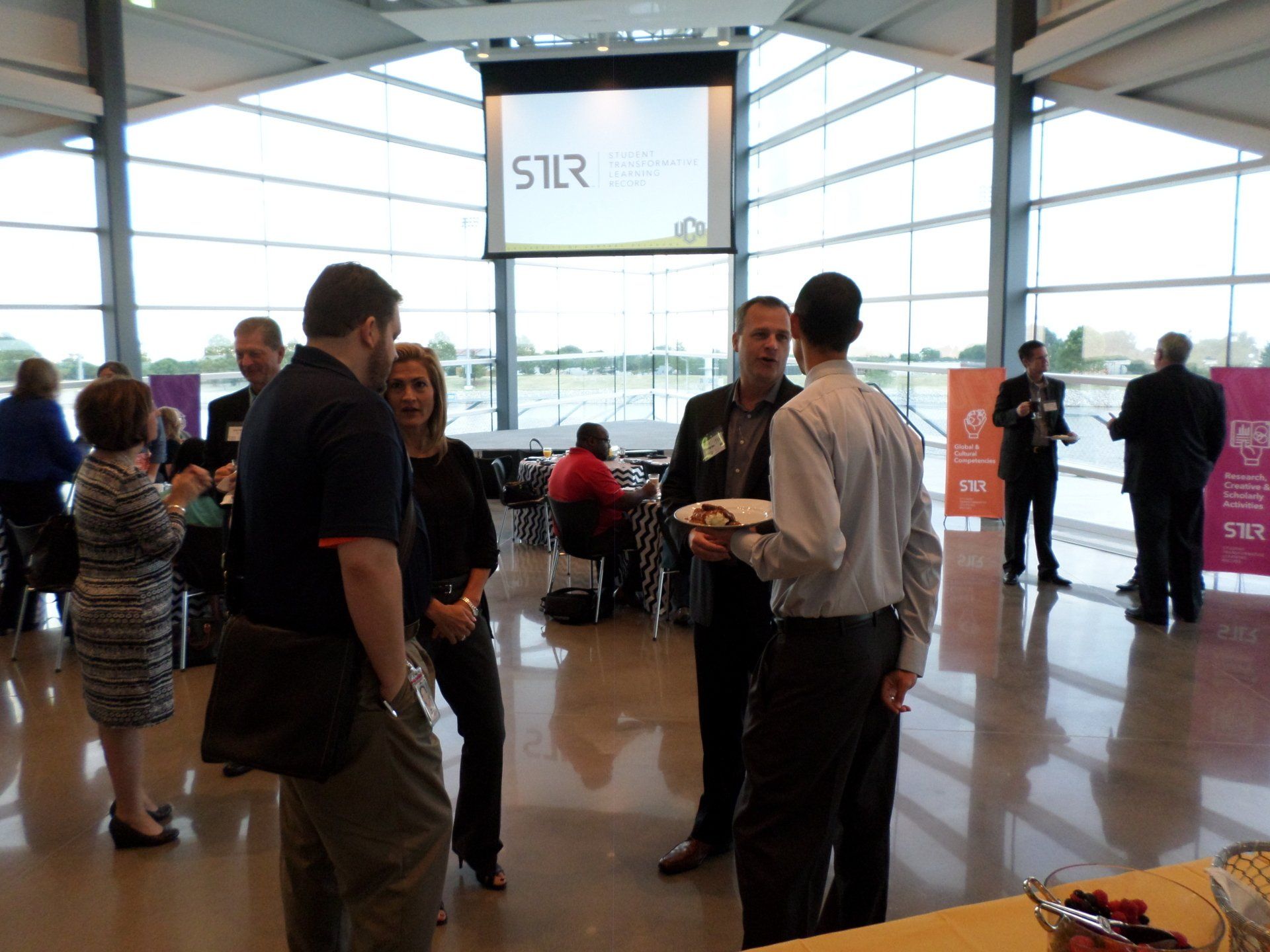 People networking at an event. Modern building with large windows. Attendees are standing, talking, and eating.