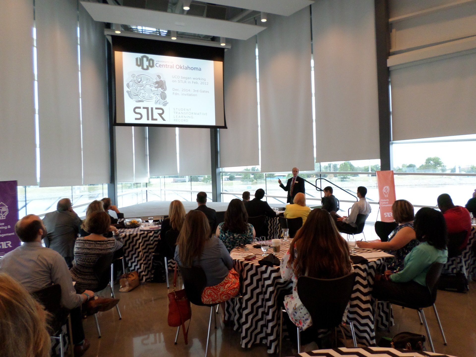People in a meeting room, listening to a presenter at a screen. Tables have black and white patterned cloths.