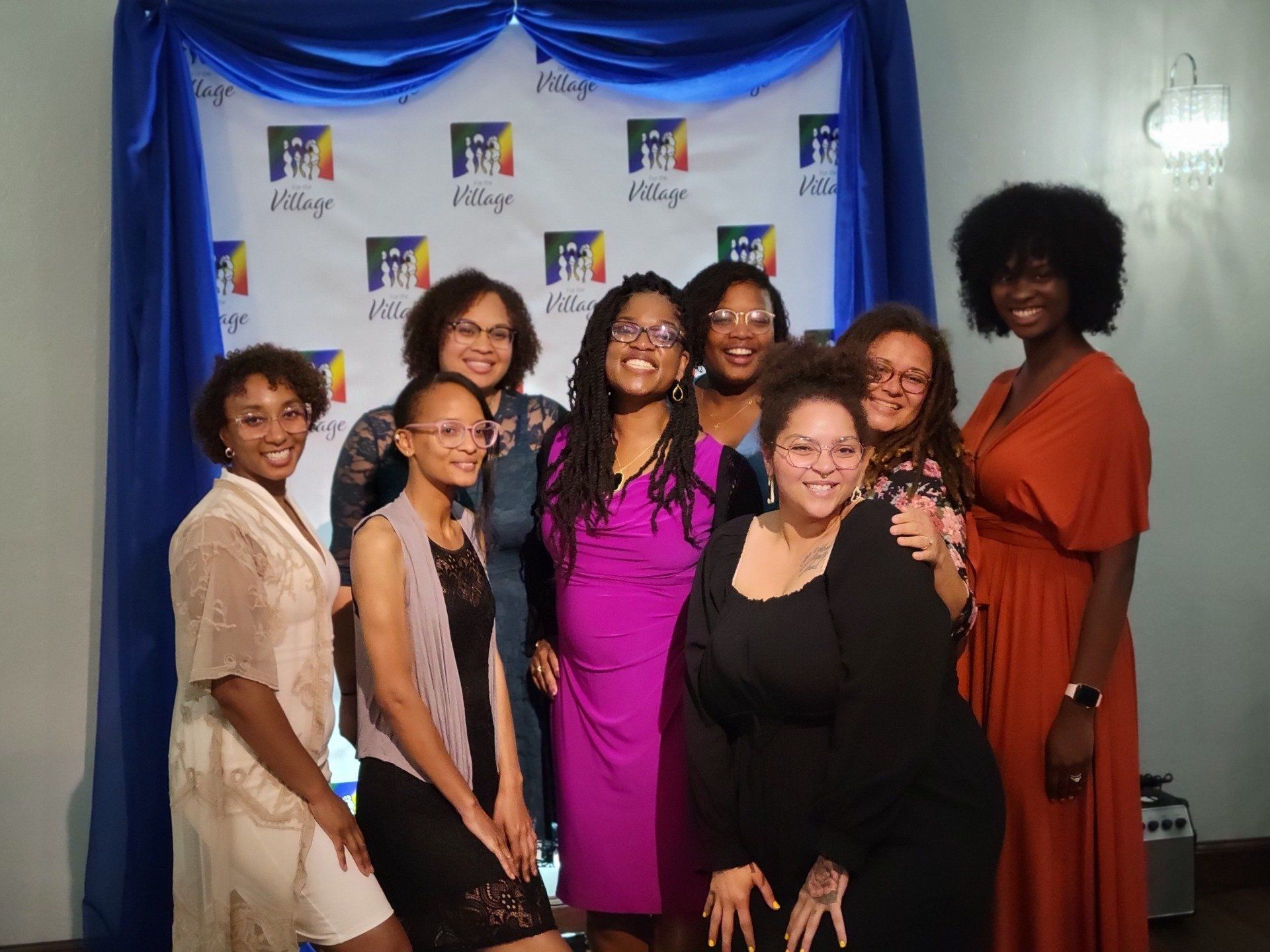 Group of women smiling in front of a blue and white backdrop with logo. Women in various dresses posing.