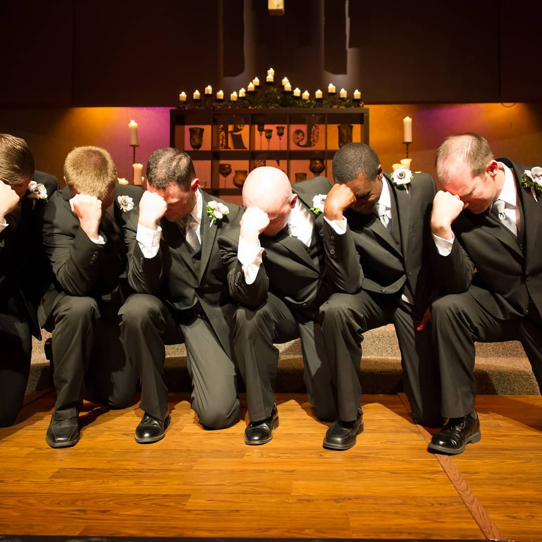 Six men in formal wear kneeling, hands on foreheads, in a dimly lit chapel before candles.