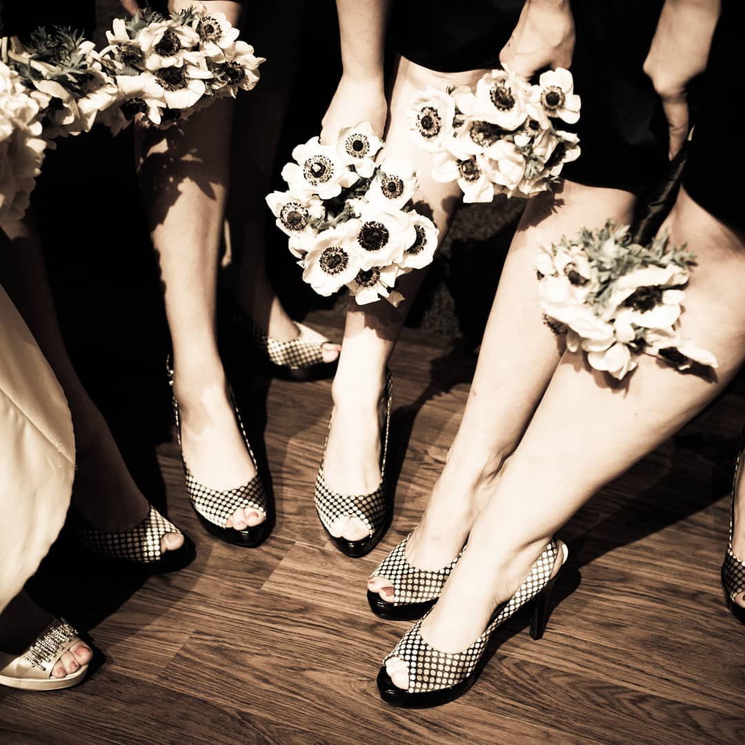 Bridesmaids holding bouquets of white flowers, wearing peep-toe checkered heels, grouped on wood floor.