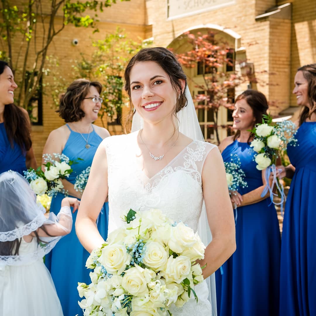 Bride in white dress, smiling, holding bouquet, surrounded by bridesmaids in blue dresses.