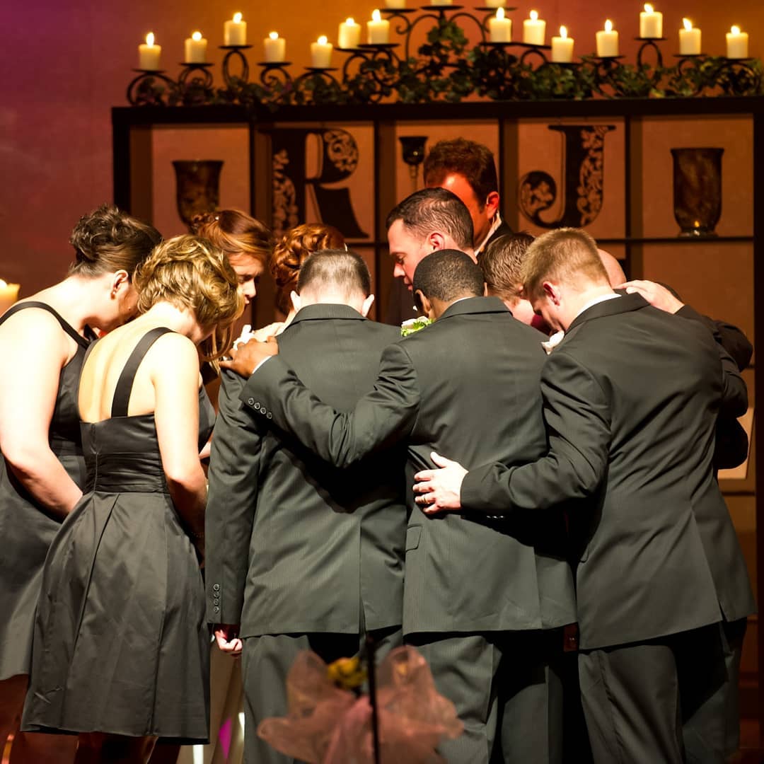 People in formal wear huddle, heads bowed, arms around each other. Candles and decorations in the background.