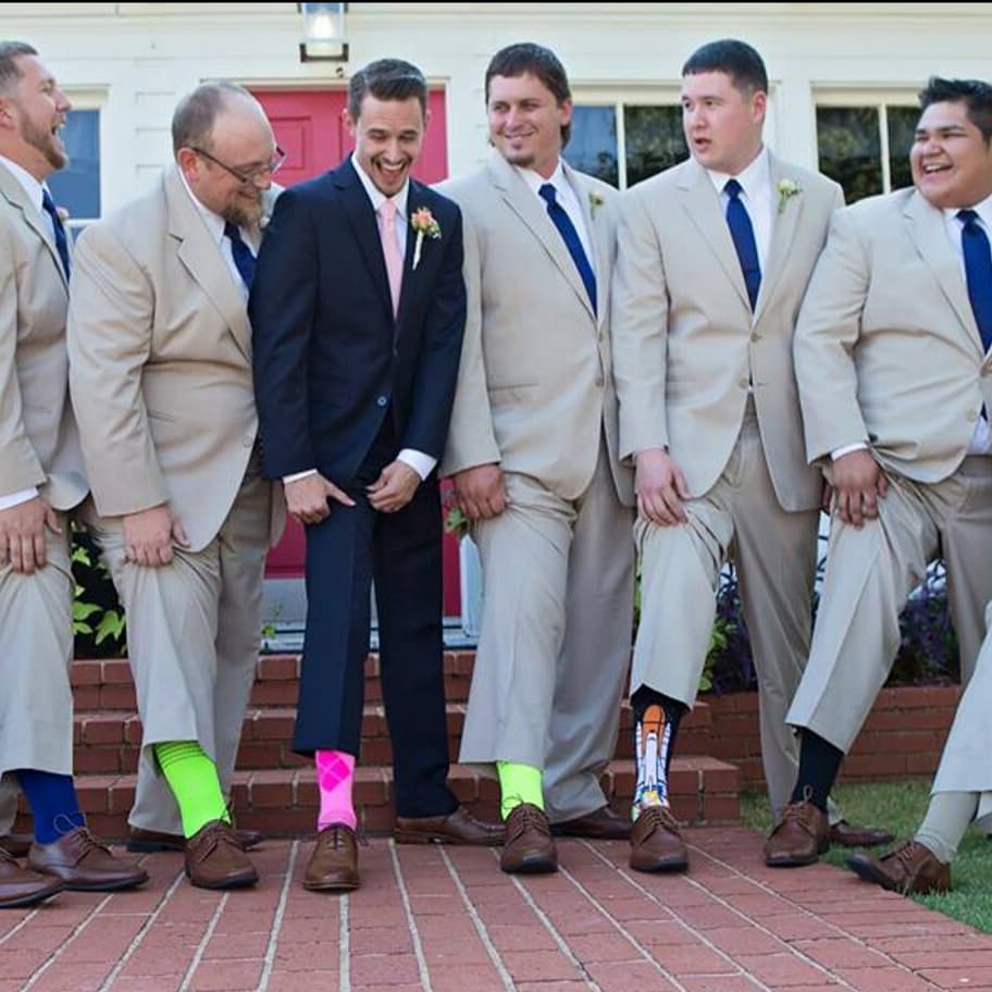 Men in suits posing, showing colorful socks outdoors. Some have hands in pockets.