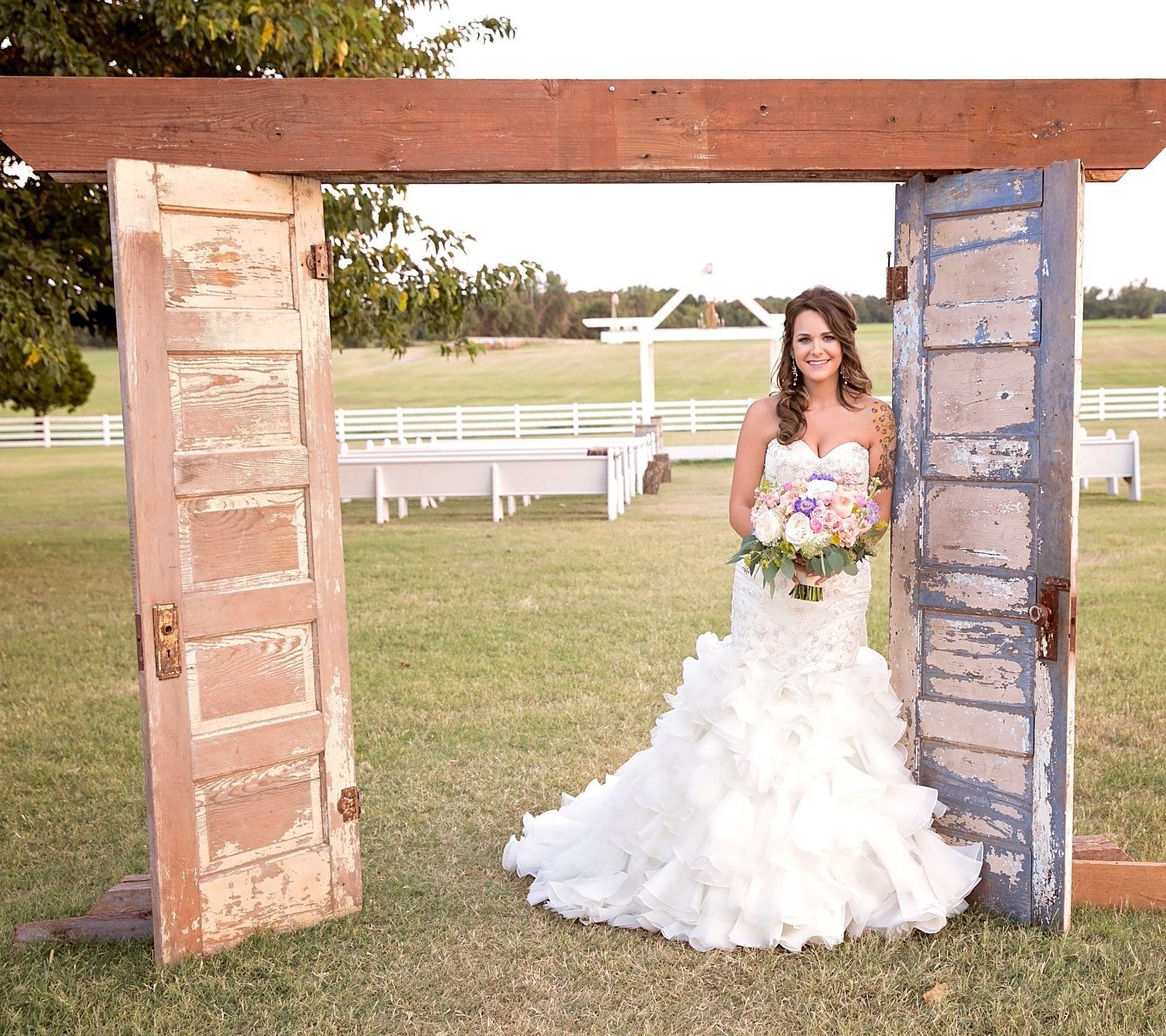 Bride in white ruffled dress poses between rustic doors, holding bouquet, in a field.