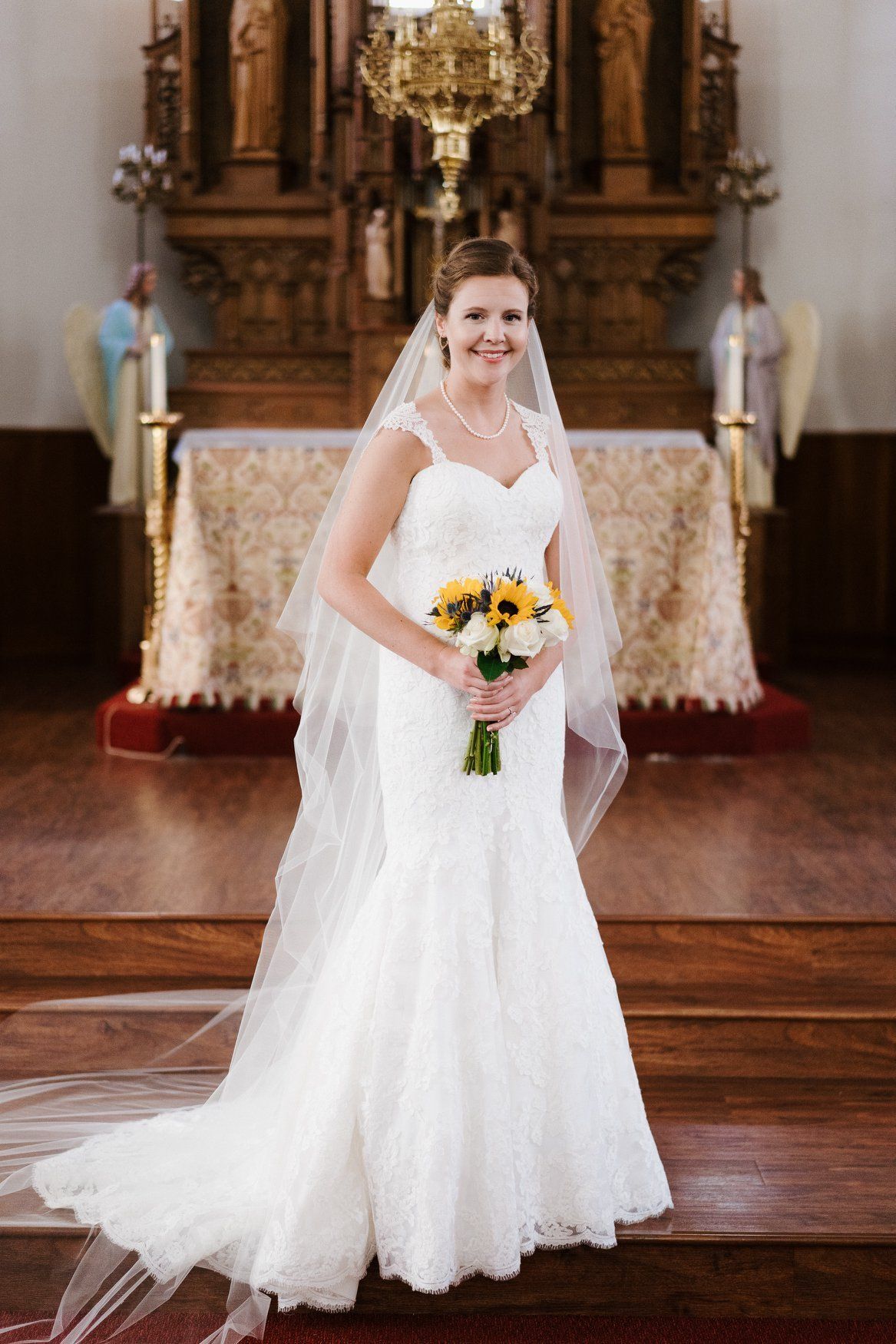 Bride in white lace dress holding sunflowers, smiling in church.