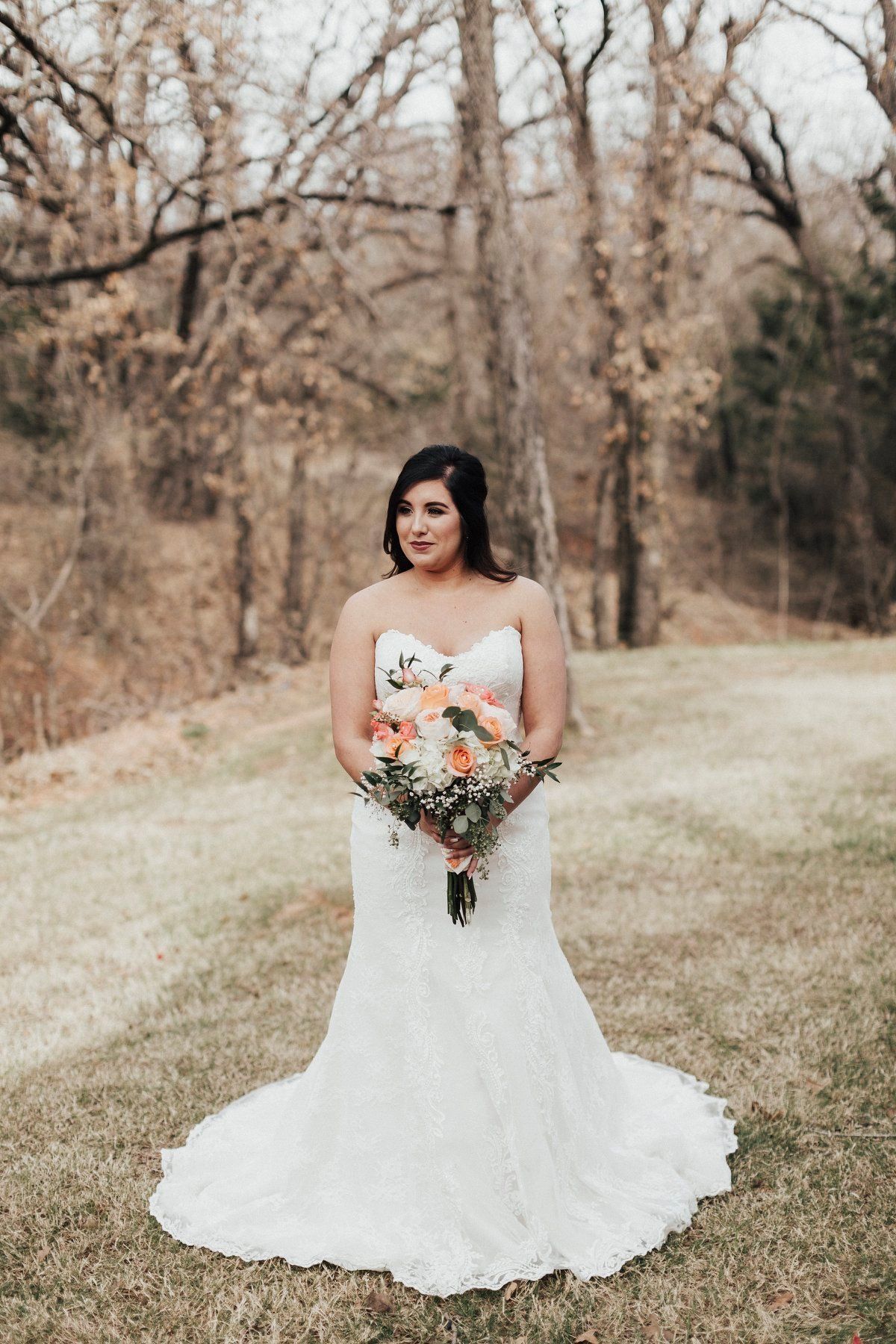 Bride in white strapless gown holds bouquet in a field with trees in the background.