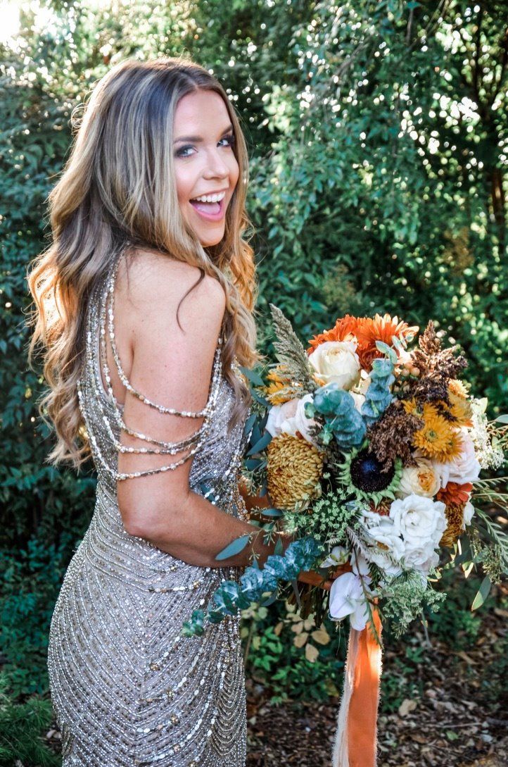 Woman in sparkly dress smiles, holding colorful bouquet. Outdoors with trees in the background.