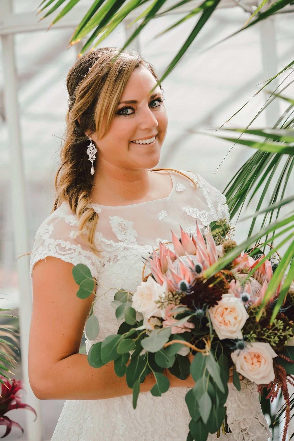 Bride in white lace dress, holding bouquet, smiling in greenhouse setting.