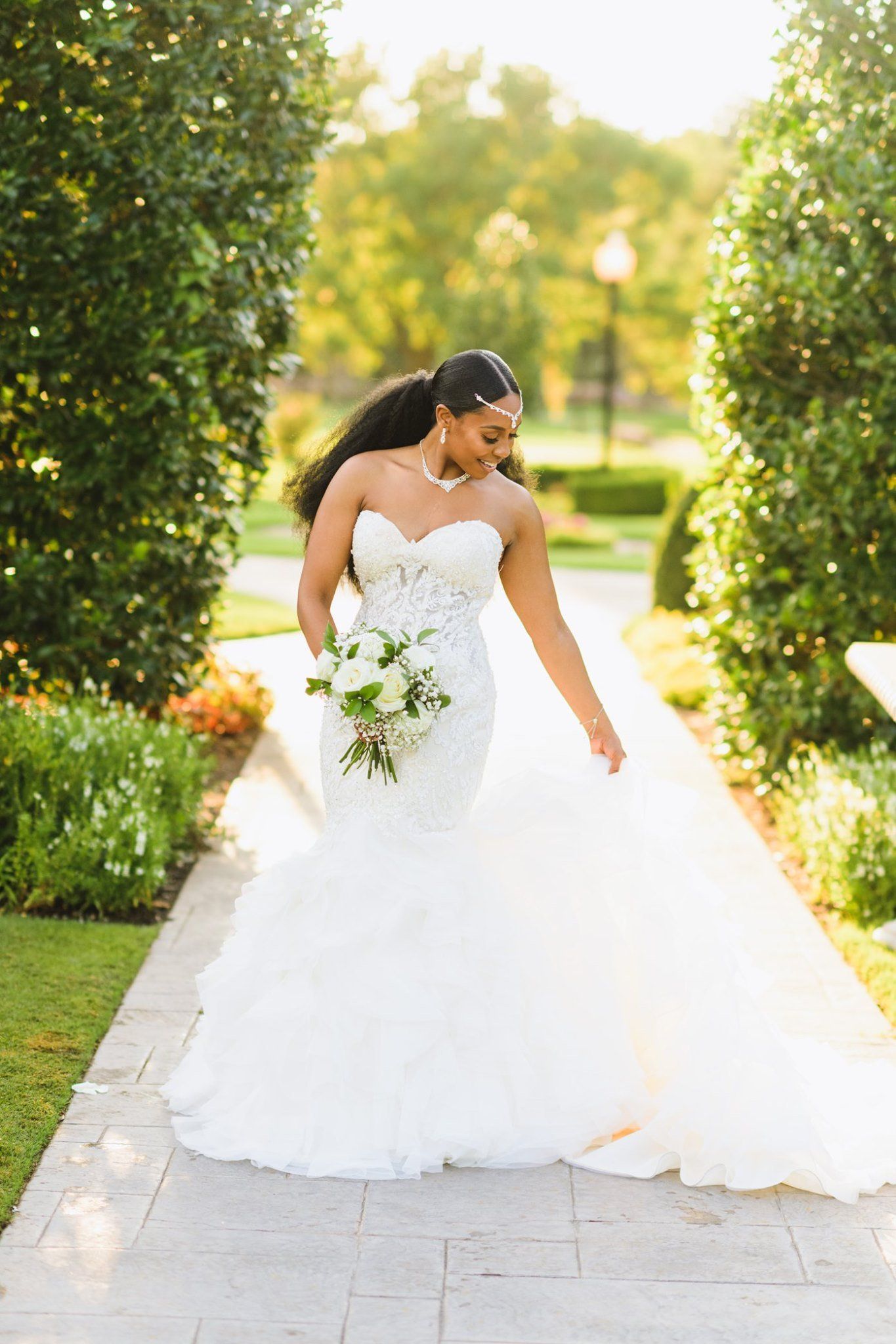 Bride in white, floral wedding dress, holding bouquet, outdoors on a stone path.