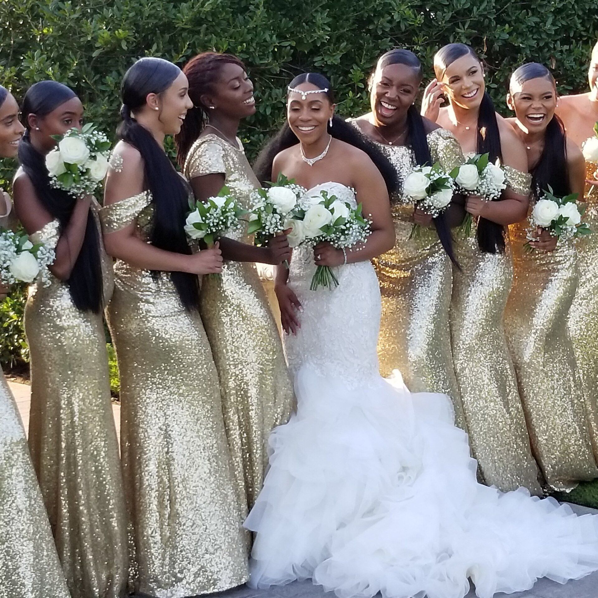 Bride in white gown with bridesmaids in gold sequin dresses, holding bouquets and smiling outdoors.