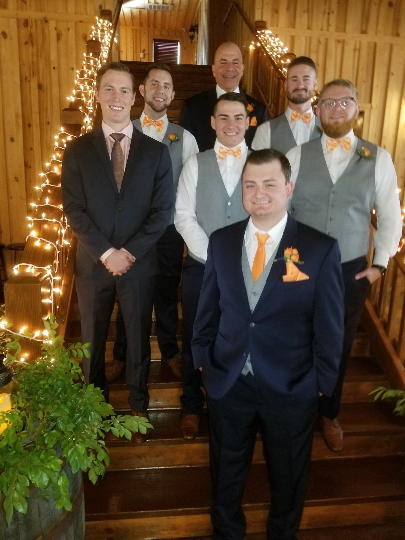 Groomsmen in suits and bow ties posing on wooden stairs decorated with lights.