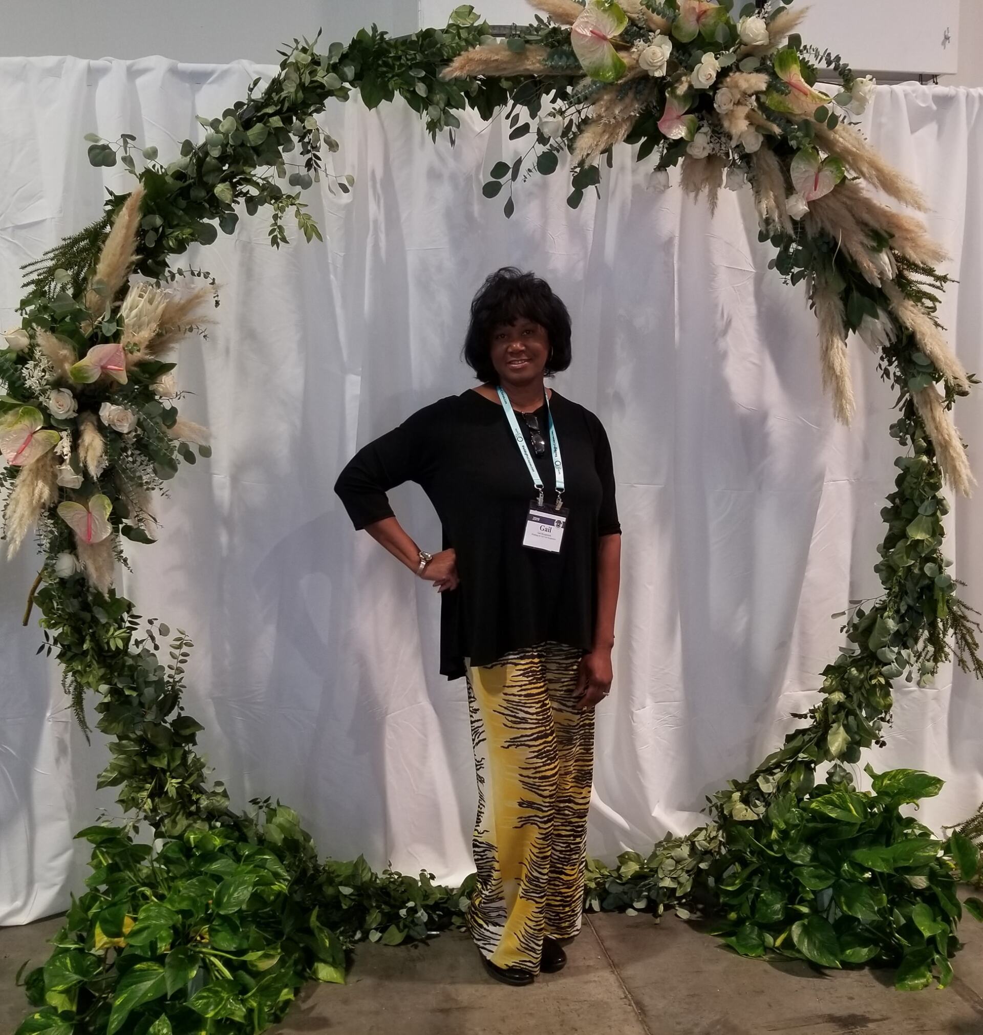 Woman posing in front of a floral archway, wearing a black top and patterned pants.