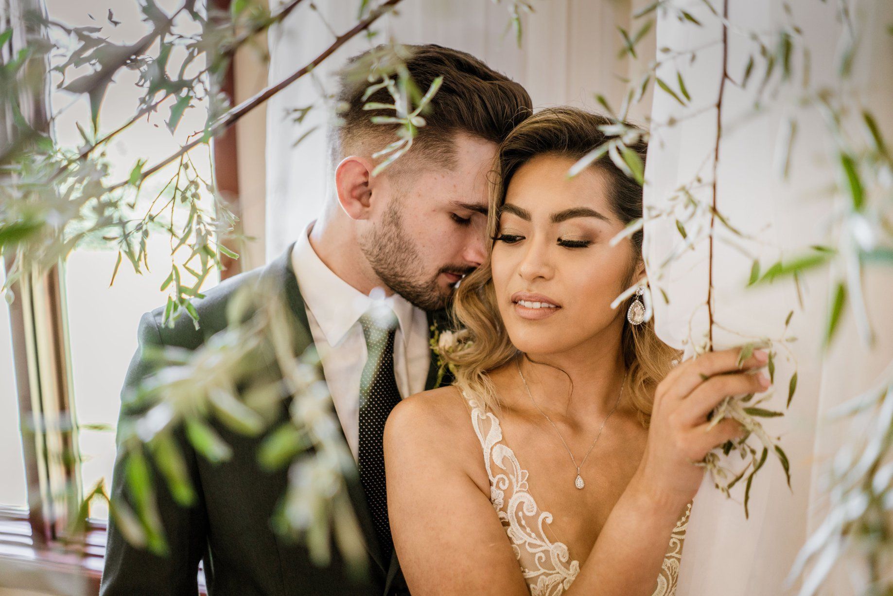 Bride and groom embrace by window, greenery framing them. Groom's head near bride's. Both have eyes closed.