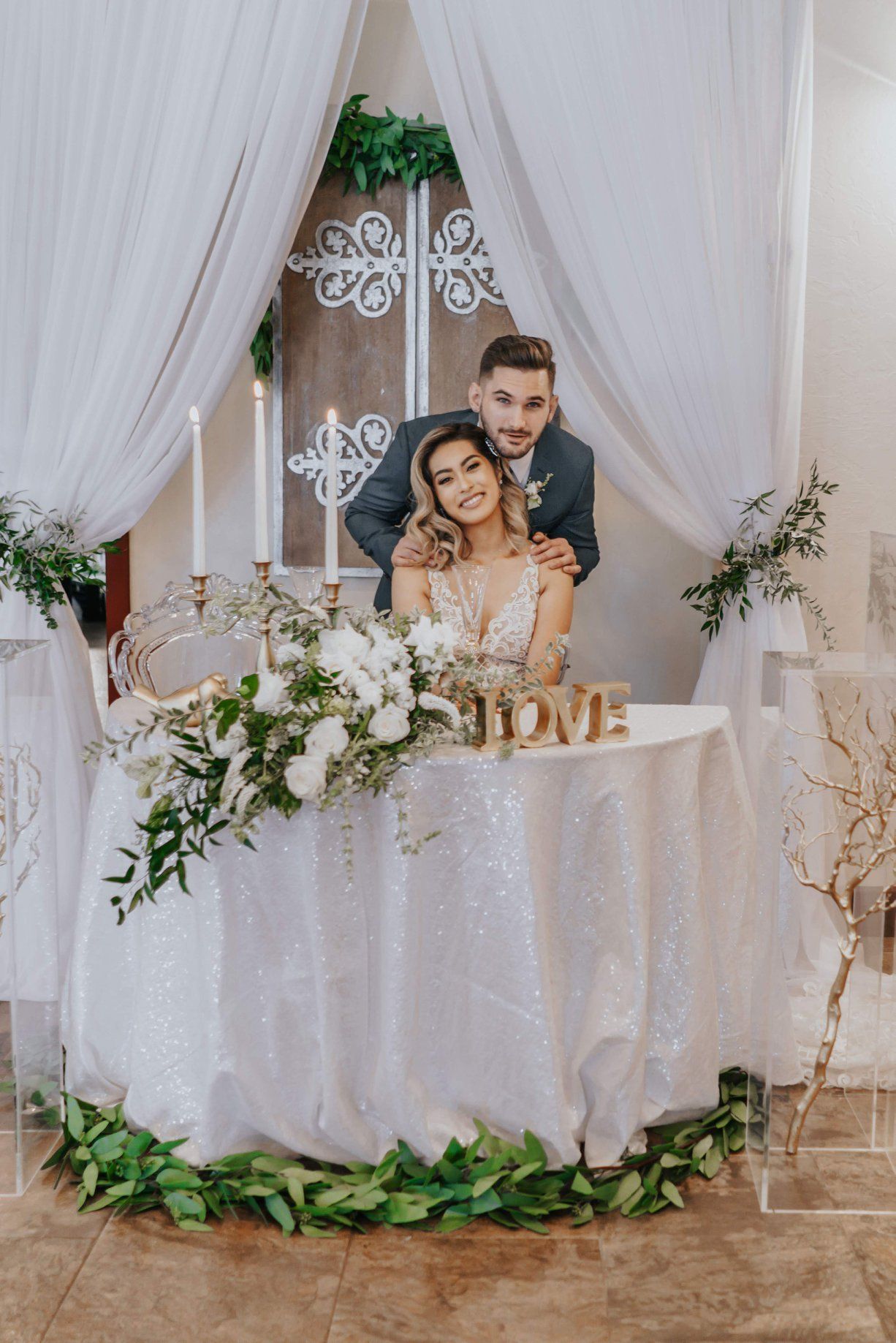 Bride and groom smiling at table decorated with flowers and greenery at wedding reception.