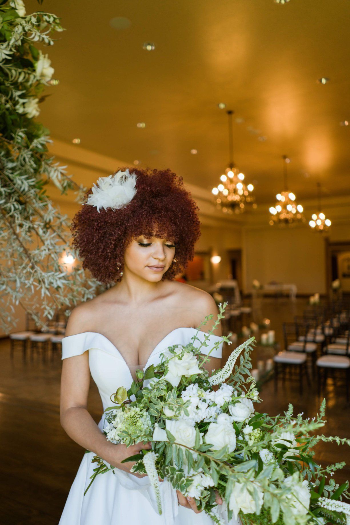 Bride in white dress holds bouquet, looking down, inside a venue with chandeliers.