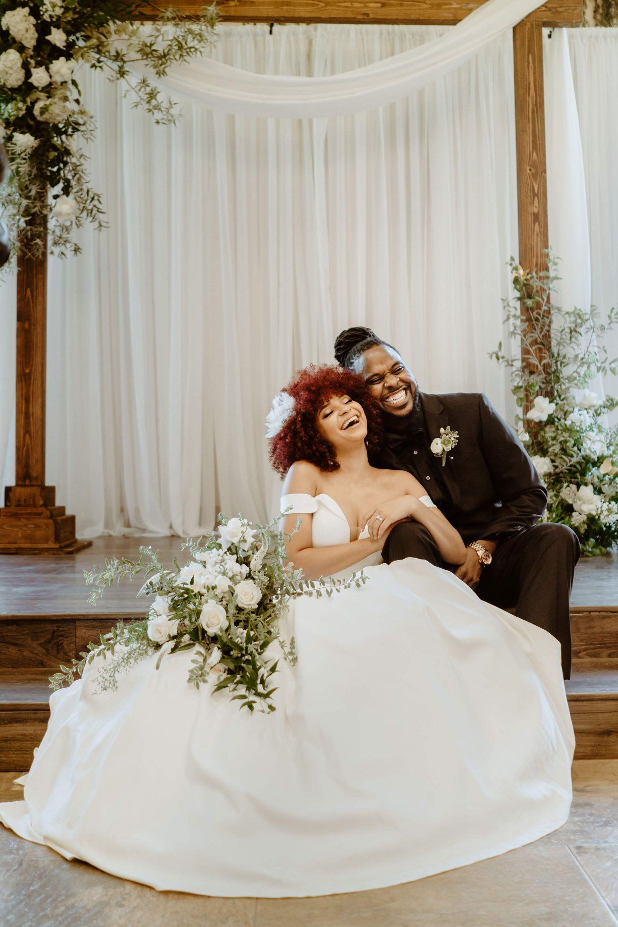 Couple laughing on a wooden platform, the bride in a white gown and the groom in a black suit, with floral decor.