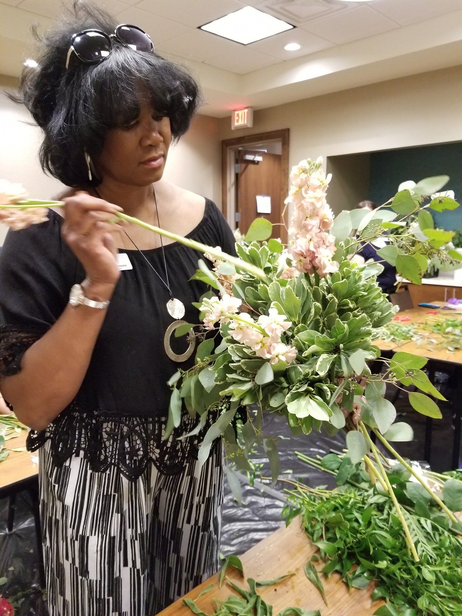 Woman arranging flowers in a workshop, holding a stem over a lush green arrangement, with floral materials on the table.