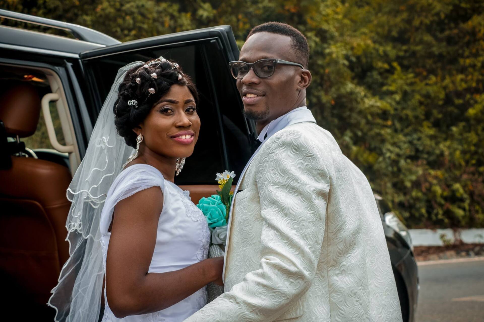 Wedding couple standing beside a black car; bride in white dress and veil, groom in white suit, smiling.