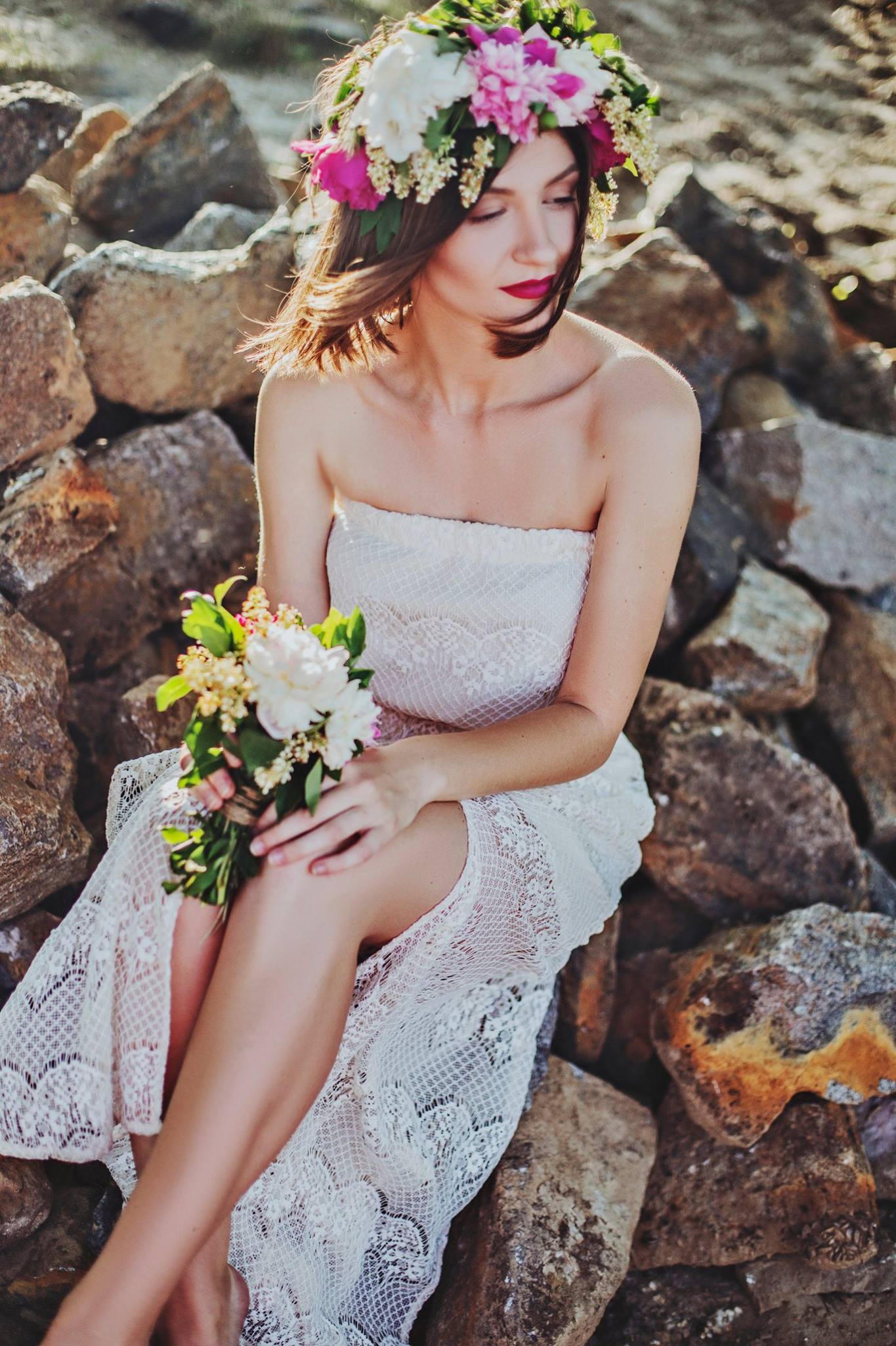 Woman in a white lace dress and flower crown sits on rocks, holding a bouquet.