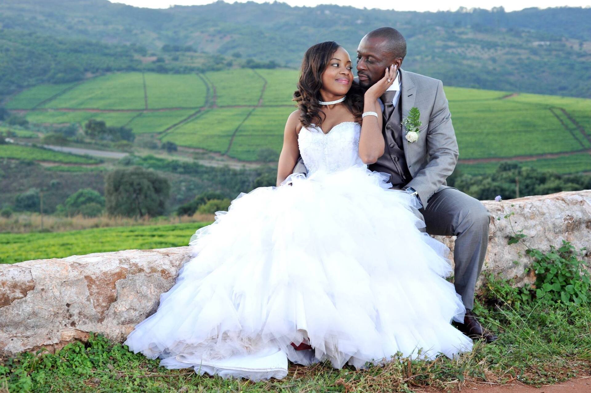 Bride and groom in wedding attire smiling, outdoors. Bride in white gown, groom in gray suit. Lush green hills in background.