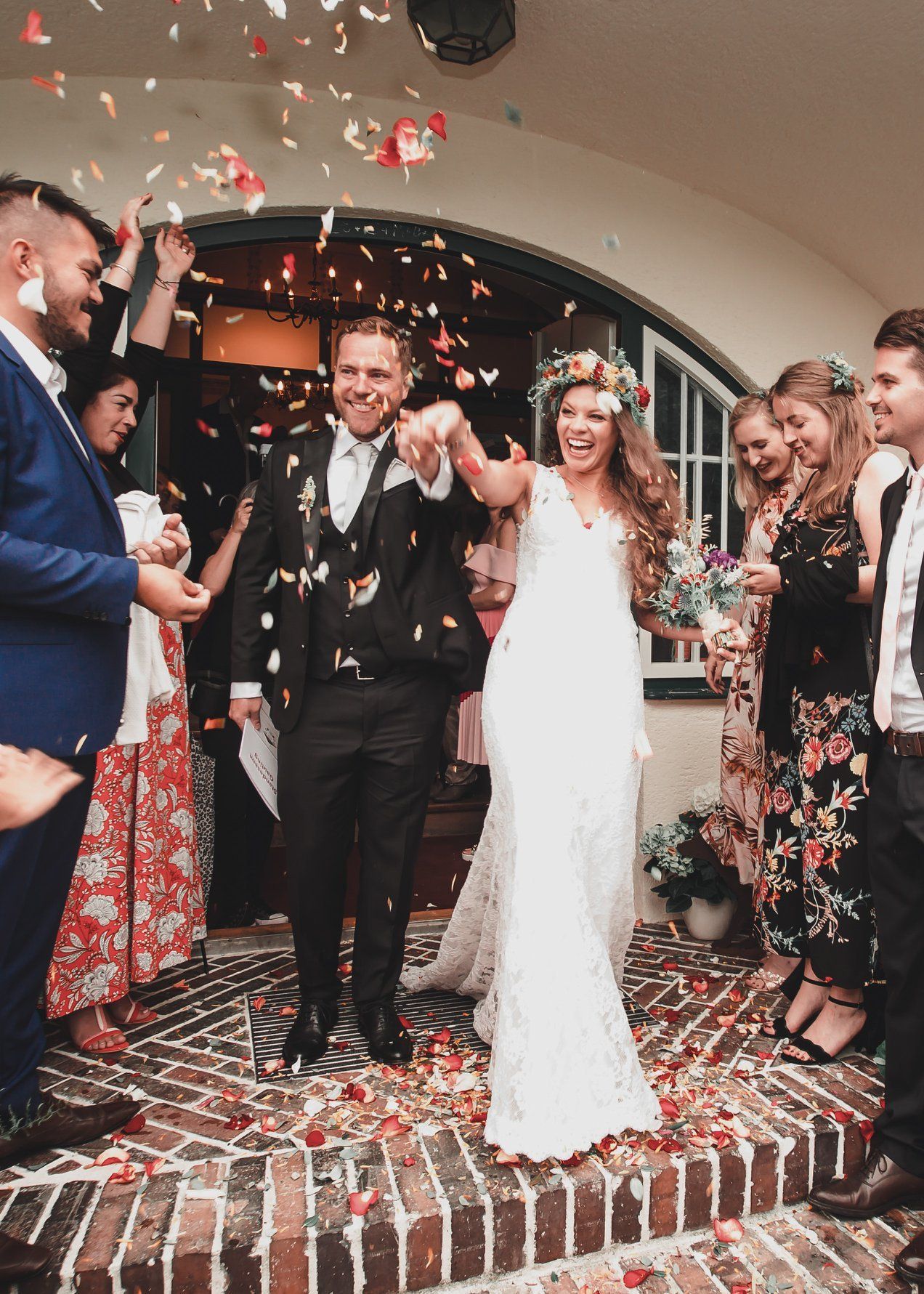 Newly married couple exits a building, confetti falling. The bride in a white dress, the groom in a suit, both smiling.