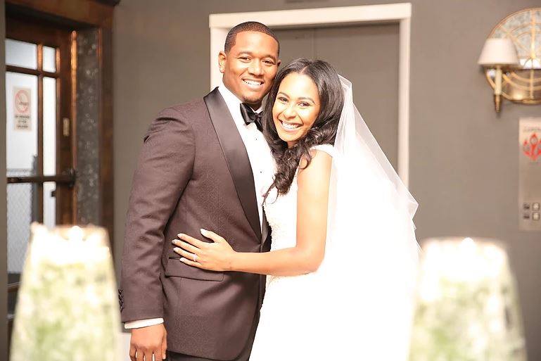 Smiling couple in wedding attire, embracing in a room with neutral walls.