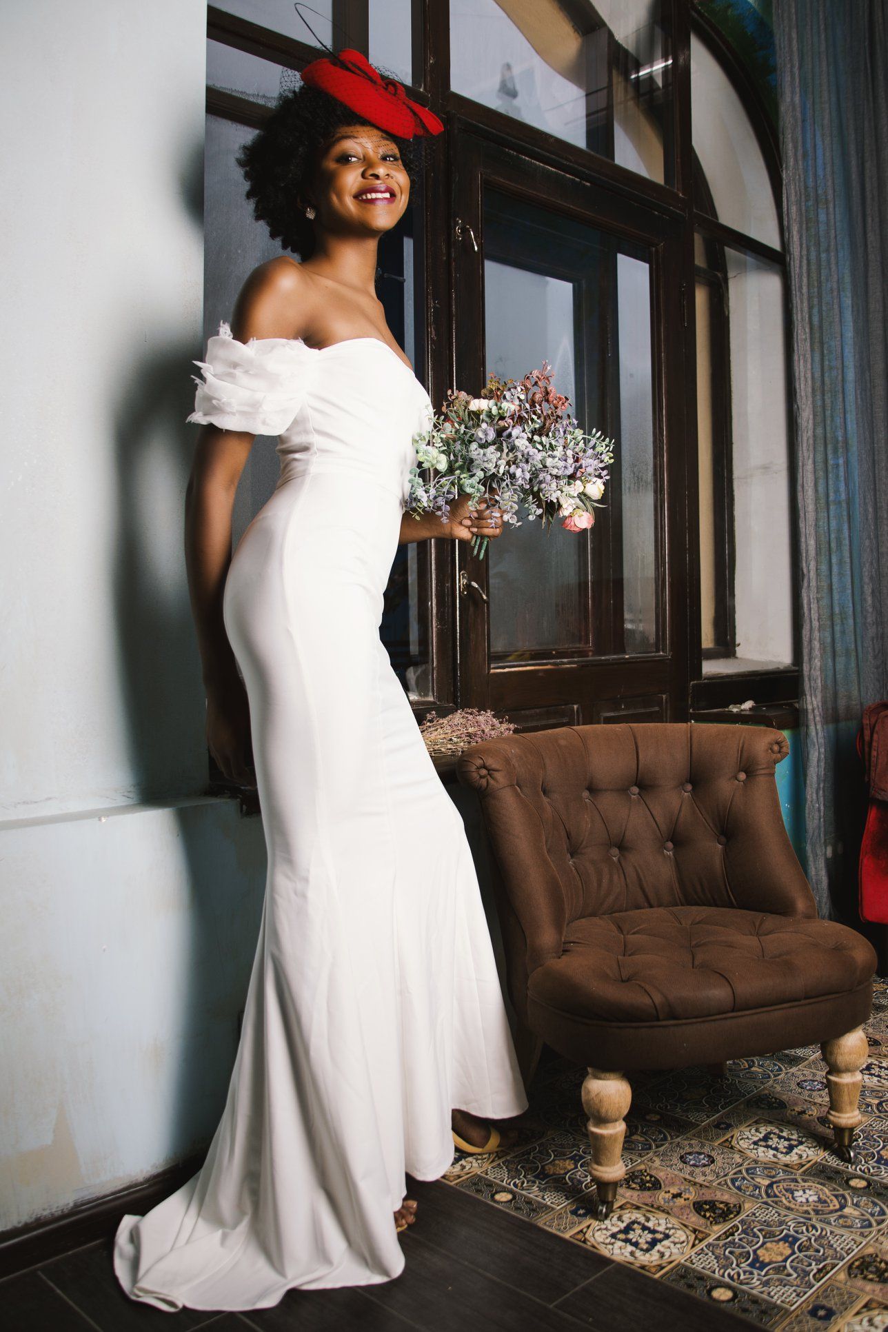 Woman in a white off-the-shoulder dress smiles, holding flowers, near a window and armchair, wearing a red hat.