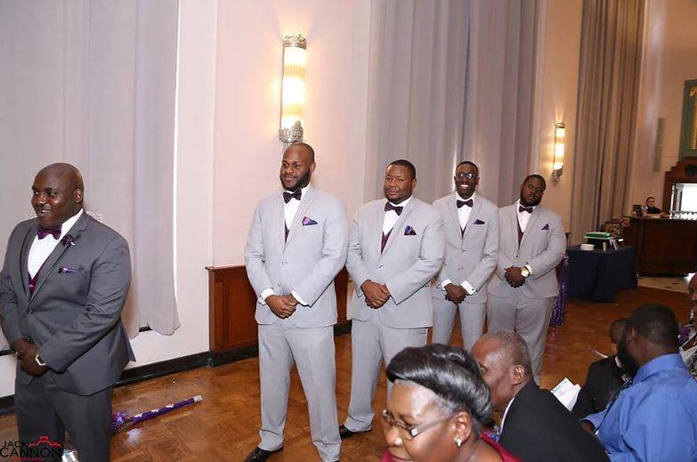 Groomsmen in gray suits at a wedding ceremony. They stand on wooden floor near an audience.