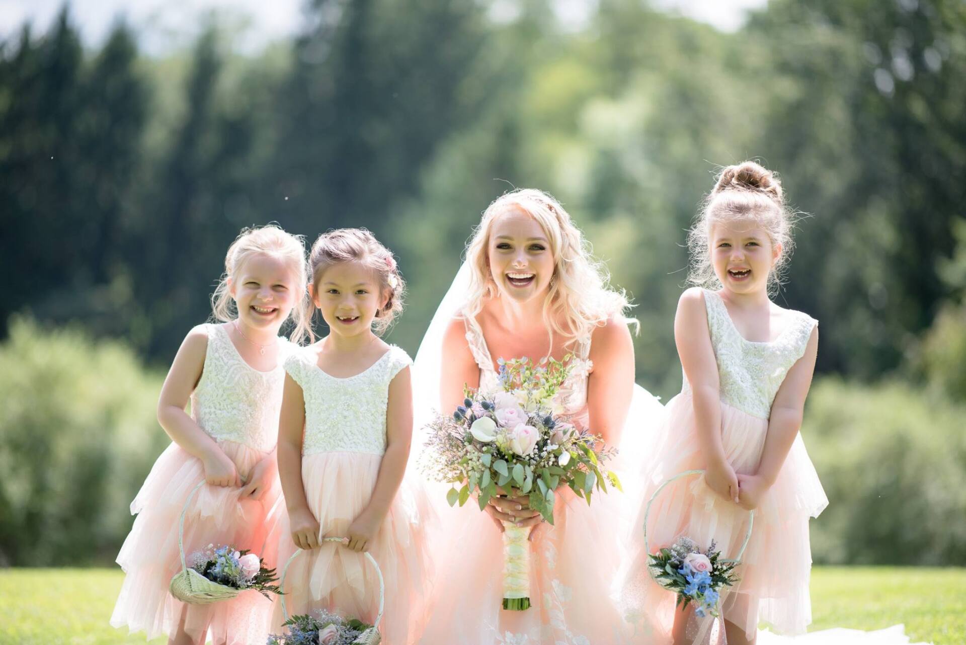 Bride with flower girls, all smiling, holding bouquets, in a sunny outdoor setting with trees in the background.