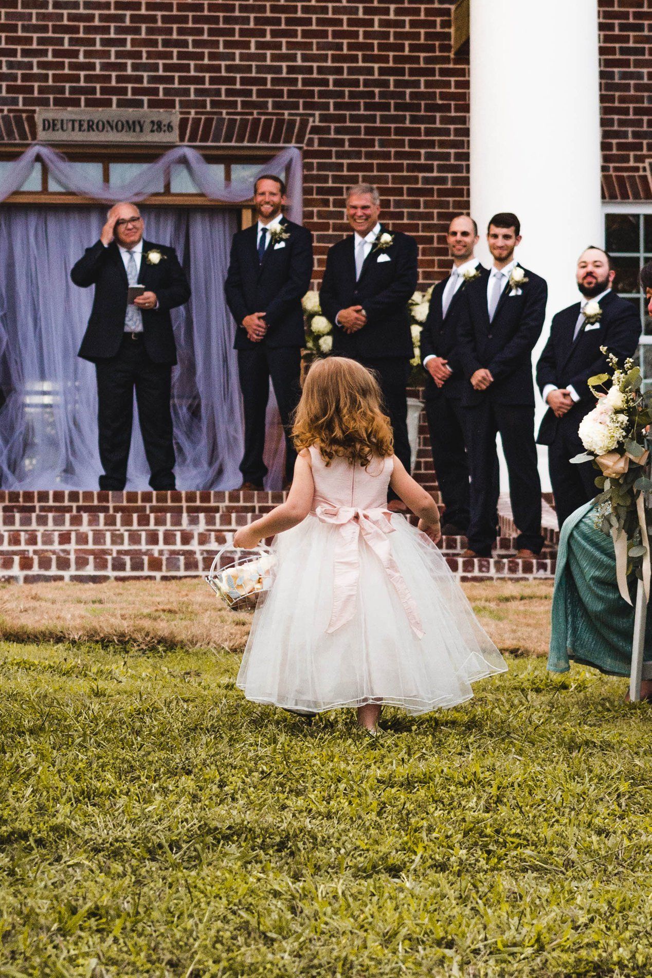 Flower girl walking toward the groom and groomsmen at a wedding ceremony on brick steps, wearing a pink and white dress.