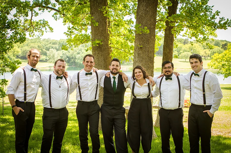 Group of people in suspenders posing outdoors near trees and a body of water.
