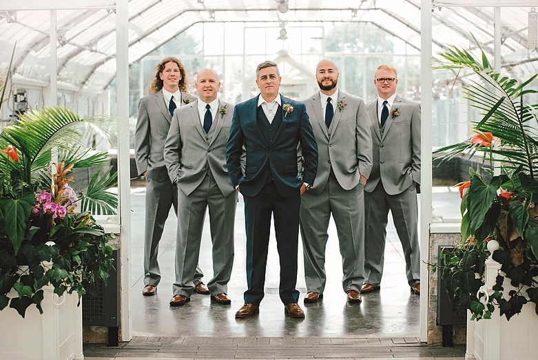 Groom in teal suit with four groomsmen in gray suits, posing in a greenhouse.