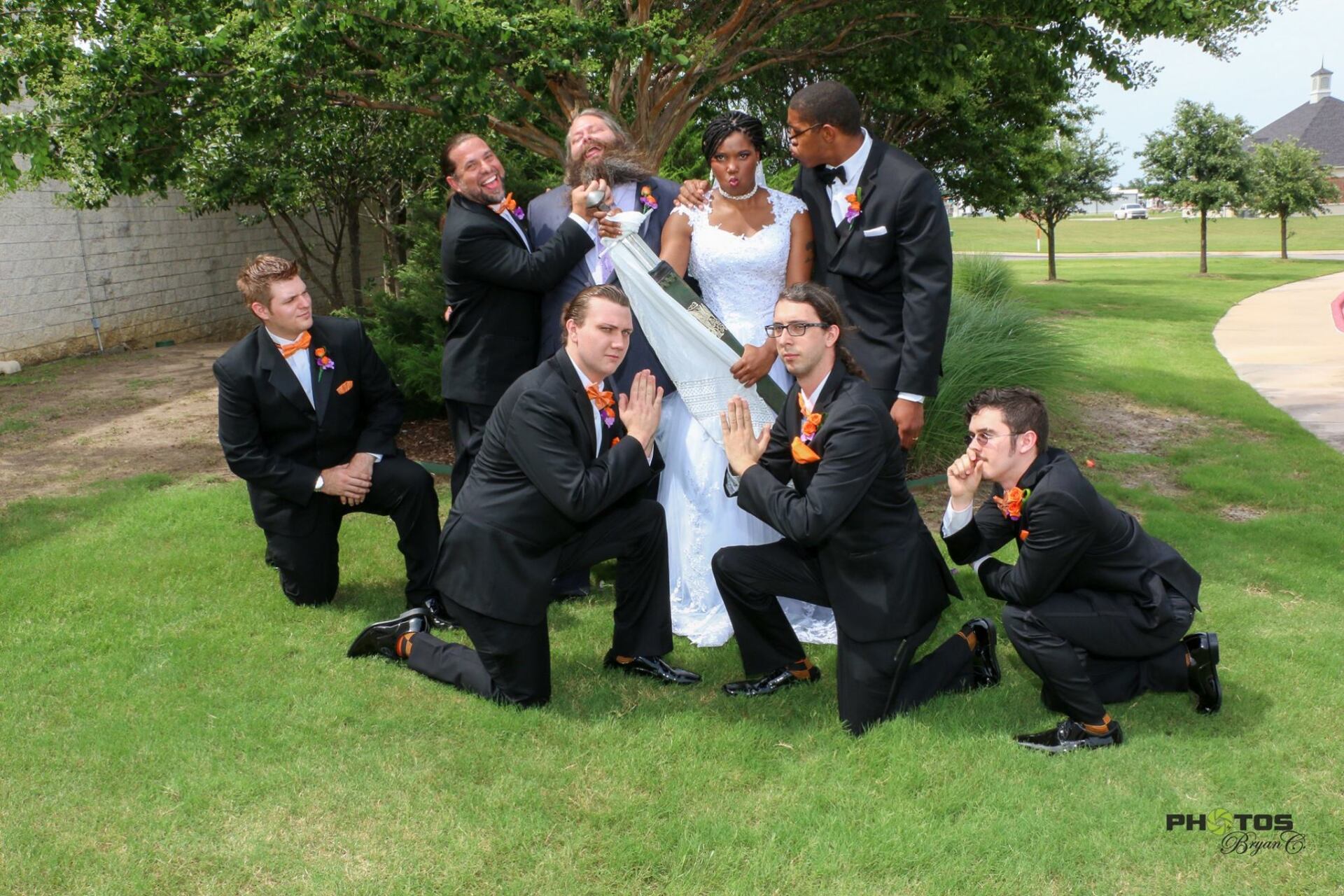 Wedding party, groomsmen surrounding bride, pulling veil, making funny faces on grass lawn.