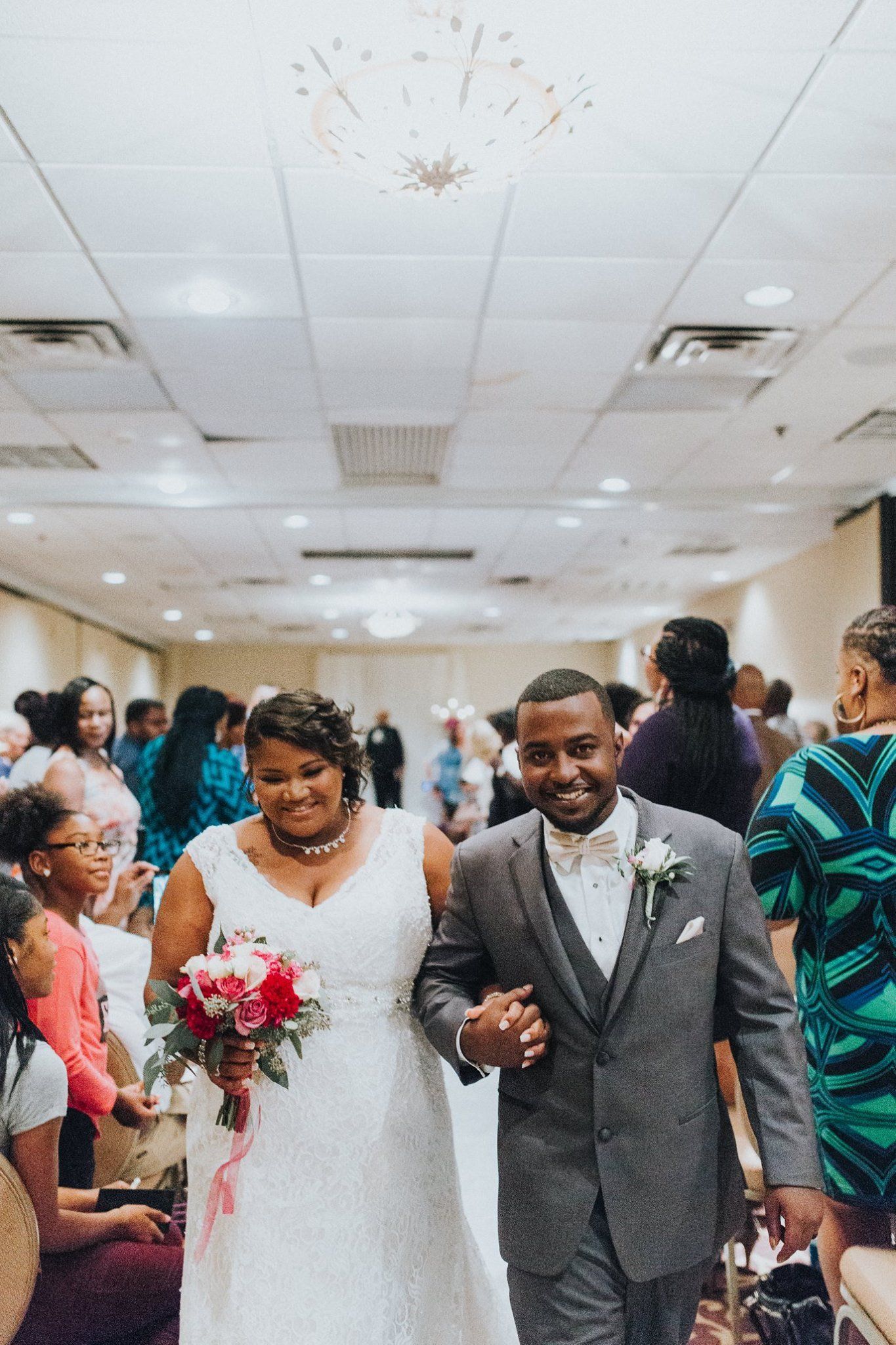 Newlywed couple walking down aisle, smiling. Bride in white dress holds bouquet. Groom in gray suit. Wedding ceremony.