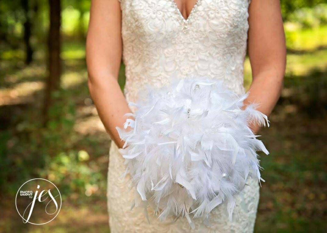 Bride in white lace dress holds a bouquet made of white feathers outdoors.