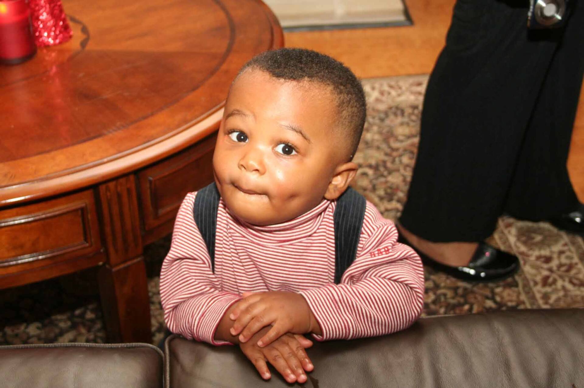 Child with dimples leans on furniture, looking at the camera.