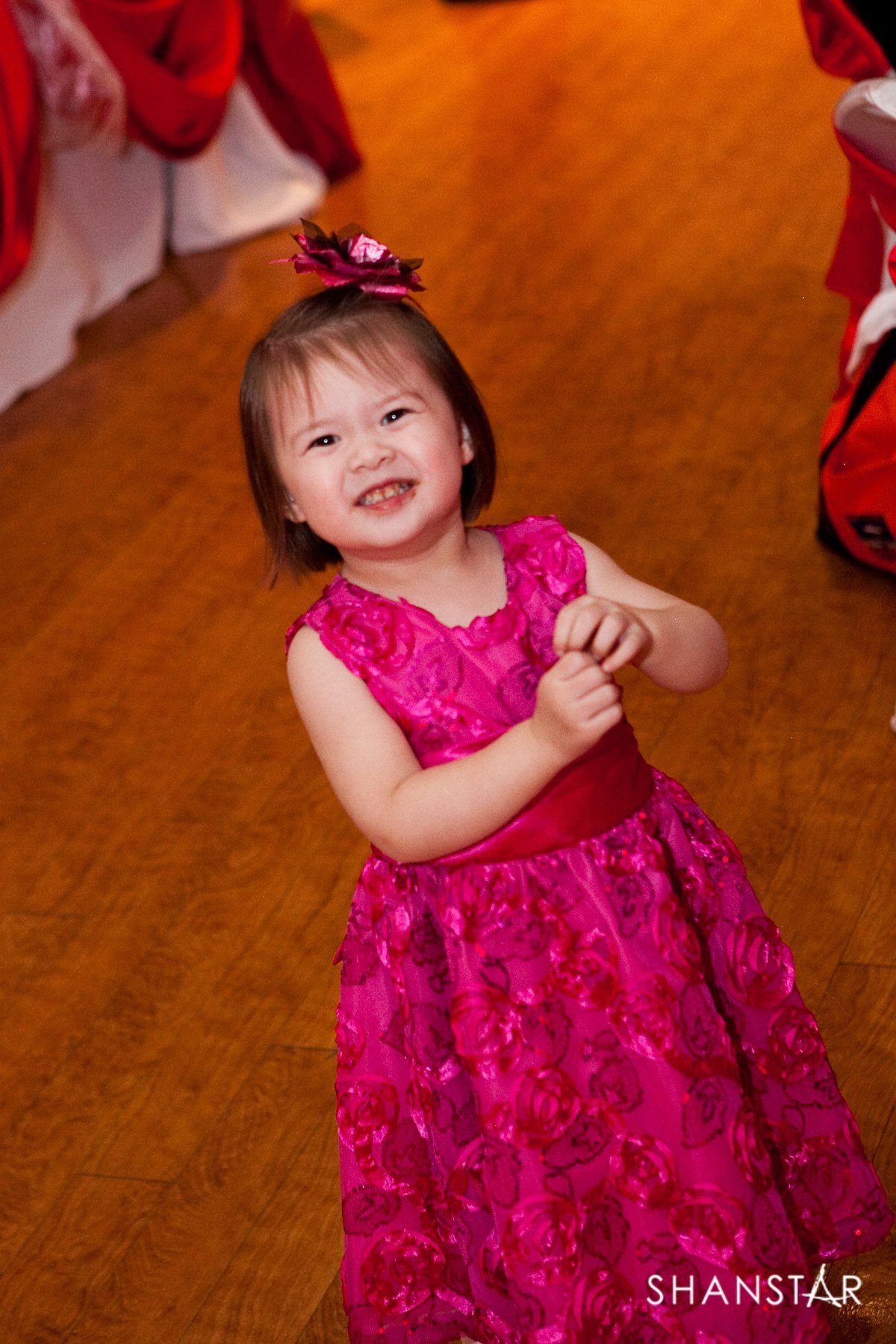 Smiling child in a pink dress and hair bow on a wooden floor, near red draped fabric.