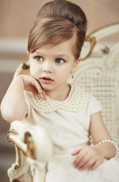 Young child with styled hair and pearl jewelry, dressed in a white dress, sits posing thoughtfully.