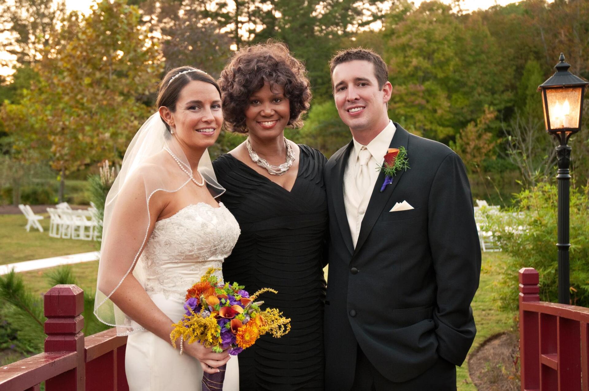 Bride and groom with a smiling officiant on a bridge, holding flowers, wedding setting.