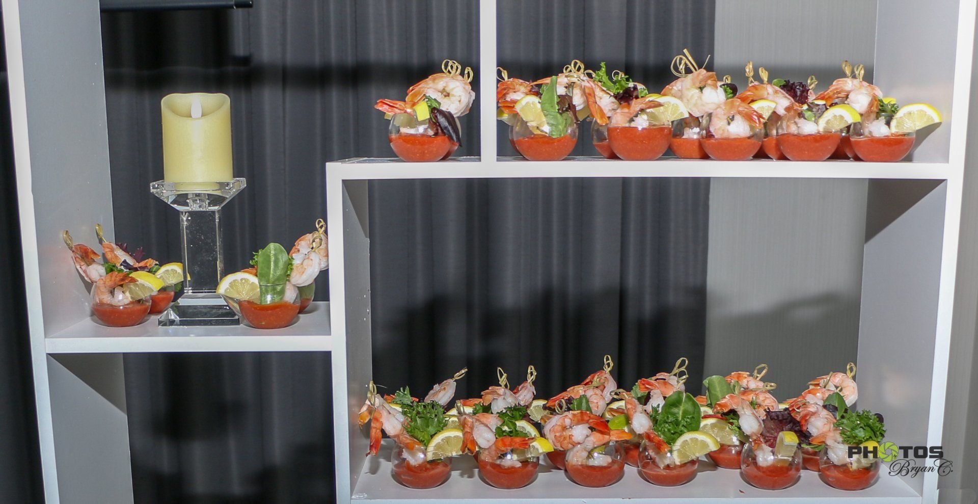 Shrimp cocktail appetizers arranged on a white shelf. Red cups with shrimp, garnish, and lemon. Candle to the left.