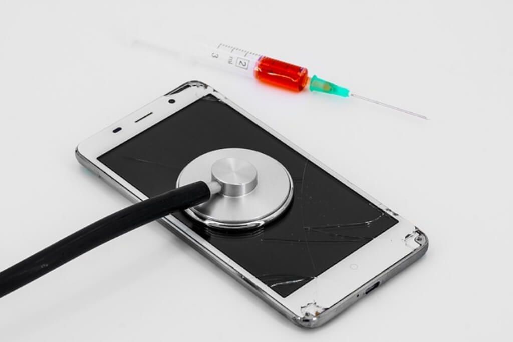 A cell phone with a broken screen is being examined by a stethoscope and syringe.